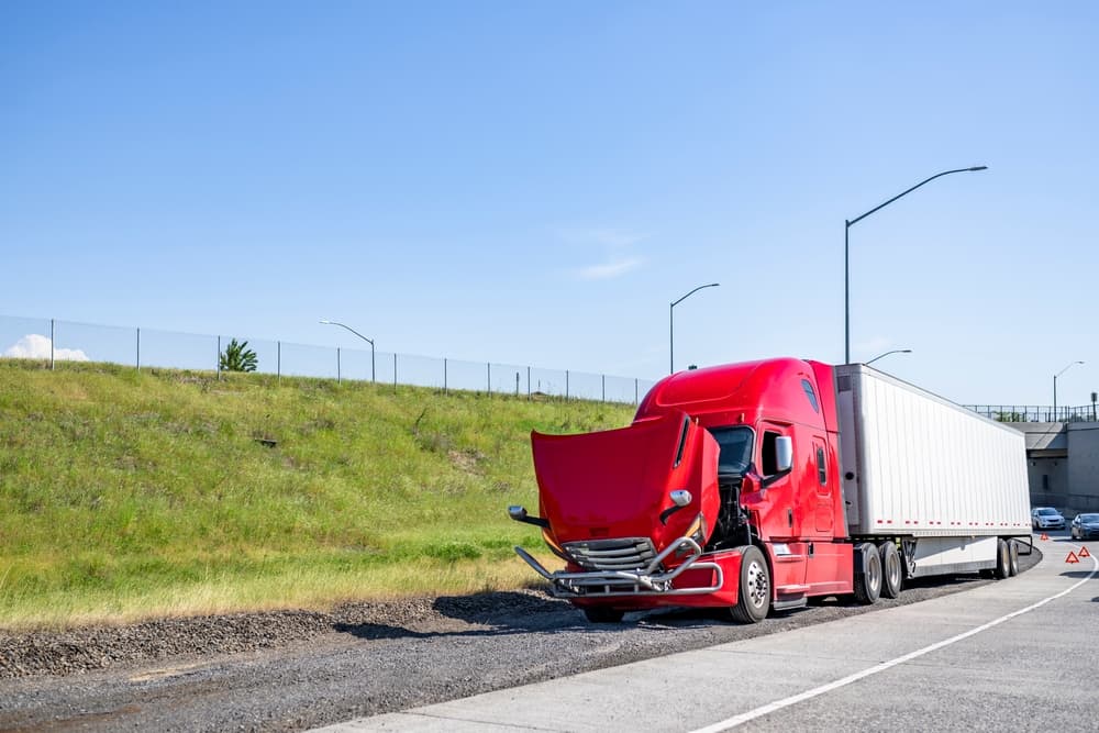 A broken red big rig semi-truck with its hood open and dry van trailer stands out of service at the highway entrance.