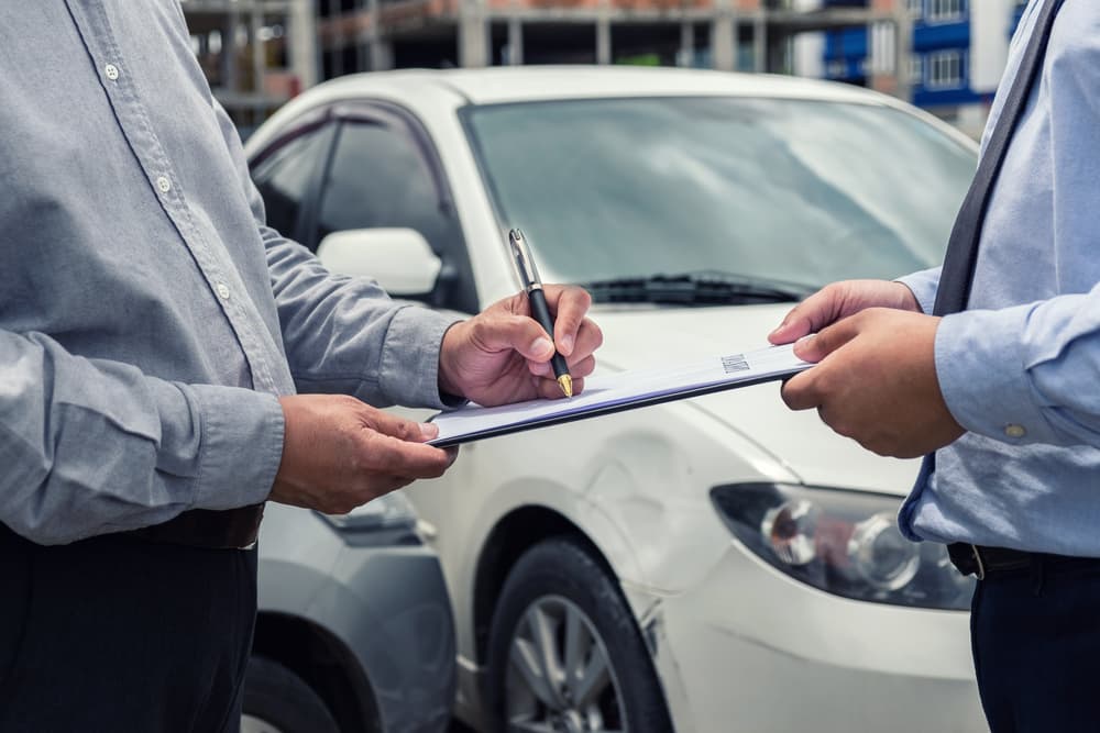 An insurance agent inspects a damaged car while the customer signs a report claim form following a traffic accident, emphasizing the claim process and insurance responsibilities.