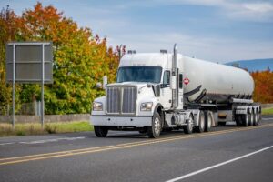 A classic industrial white bonnet semi-truck tractor hauling a specialized high-pressure armored tank trailer filled with hazardous liquefied gas, driving on the road.